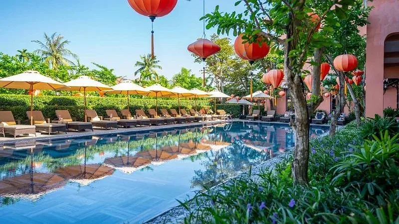 Hồ bơi dài ở Allegro Hoi An với hàng dù và ghế nằm, đèn lồng đỏ treo trên cành cây, tường hồng bên phải