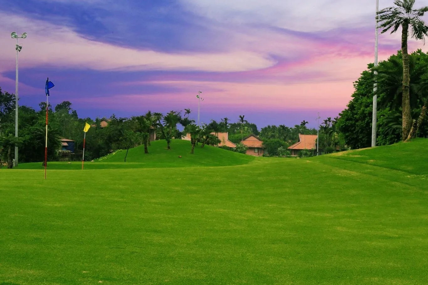Sân Asean Golf 24h tại Asean Resort & Spa: thảm cỏ xanh, cờ hố và cột đèn chiếu sáng, phía sau là mái ngói đỏ lúc hoàng hôn
