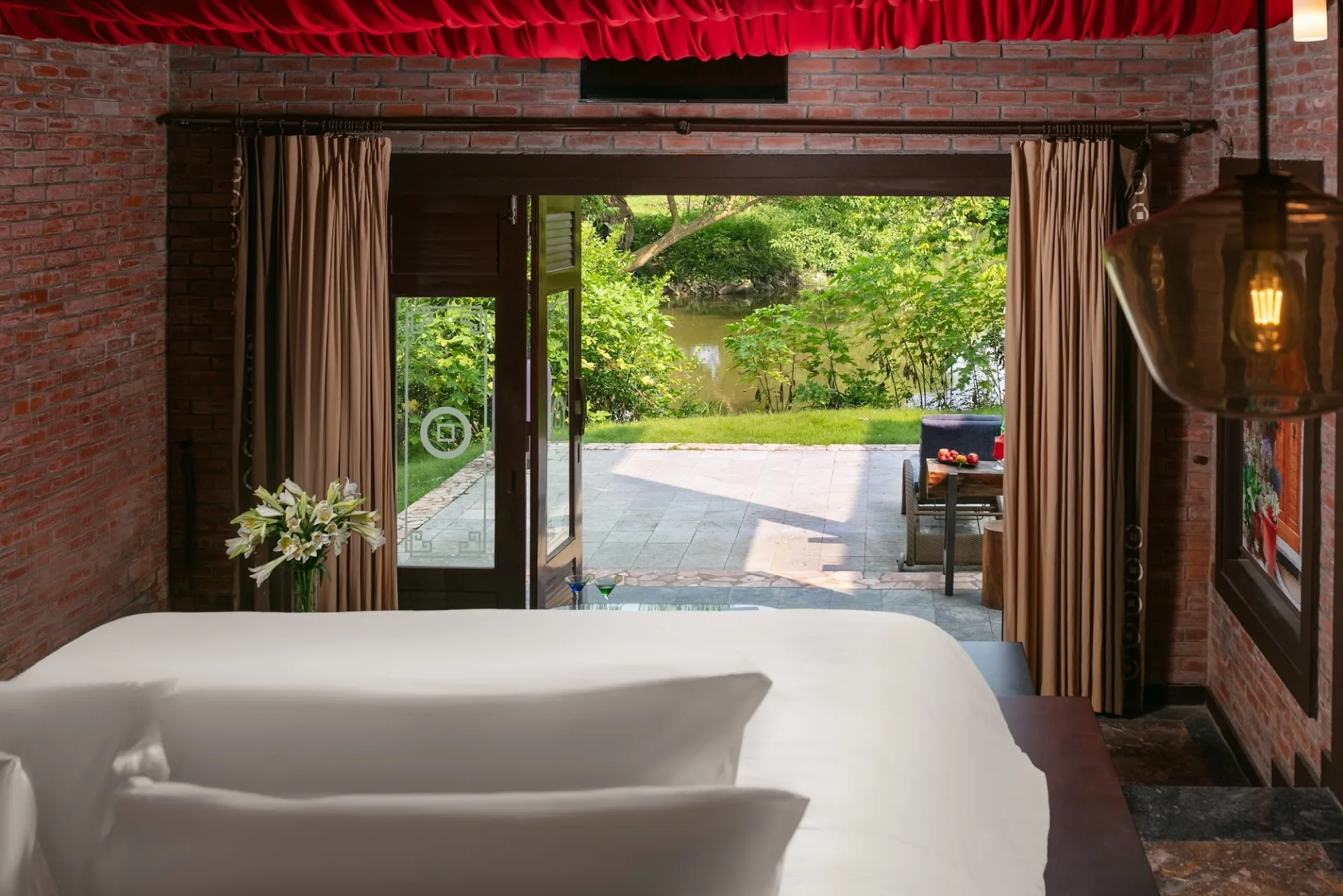 Nhìn từ giường Asean Grand Bungalow qua cửa lớn ra hiên lát đá, bãi cỏ và dòng suối trong khuôn viên Asean Resort