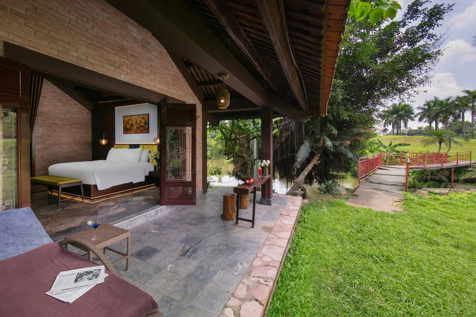 Hiên bungalow ở Asean Resort mở ra bãi cỏ, cầu gỗ lan can đỏ bắc qua suối và hàng cọ