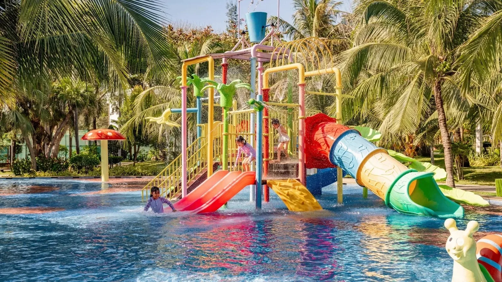 Công viên nước trẻ em tại Carmelina Beach Resort Ho Tram với cầu trượt ống xanh vàng, khung leo nhiều màu và nấm phun nước giữa hàng dừa