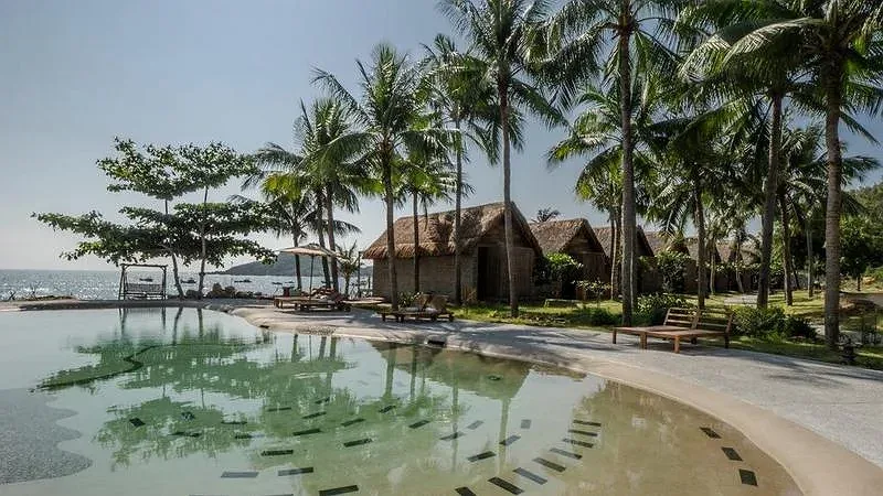 Hồ bơi uốn lượn giữa hàng dừa của Casa Marina Resort Quy Nhơn, phía sau là dãy bungalow mái lá và biển