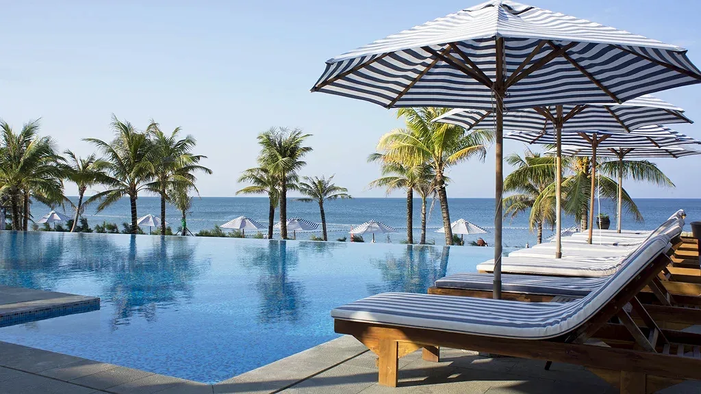 Hồ bơi vô cực của Cassia Cottage Resort Phú Quốc với ghế tắm nắng, dù sọc xanh trắng và hàng dừa nhìn ra biển