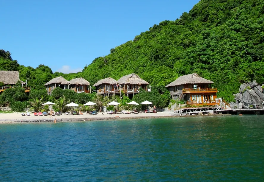Dãy bungalow gỗ mái lá của Cát Bà Beach Resort trên đảo Cát Dứa, sát bãi cát và mặt nước xanh, phía sau là đồi rừng