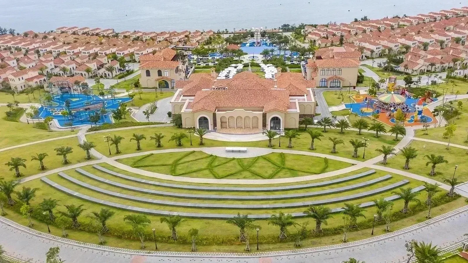 Khu Centara Mirage Resort Mui Ne nhìn từ trên cao, hai sân chơi trẻ em lớn màu xanh và màu cam nằm hai bên nhà chính mái ngói đỏ, phía sau là hồ bơi và biển