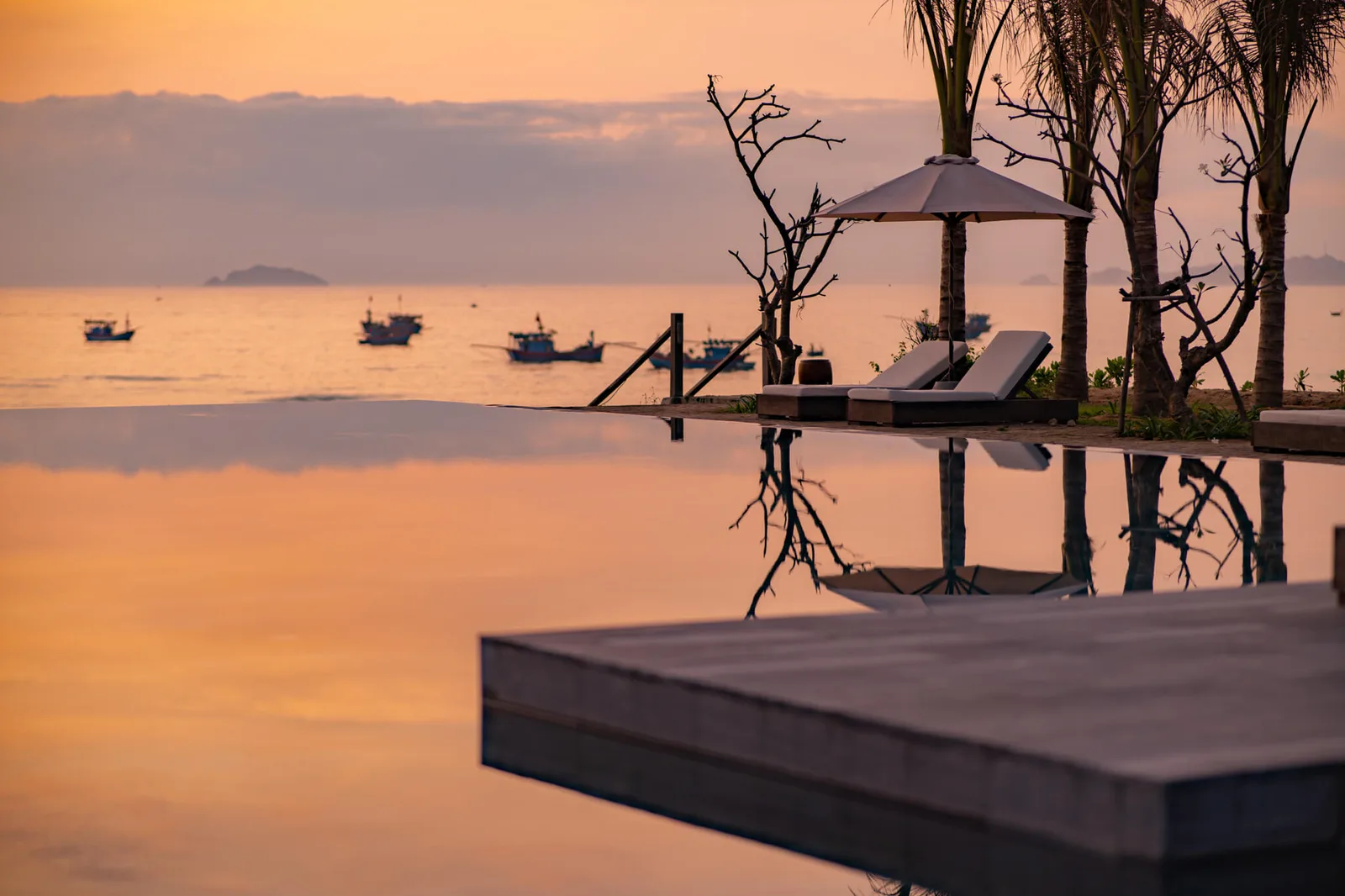 Hồ bơi vô cực của Crown Retreat Quy Nhơn lúc hoàng hôn, mặt nước phản chiếu ô dù và thuyền chài ngoài vịnh