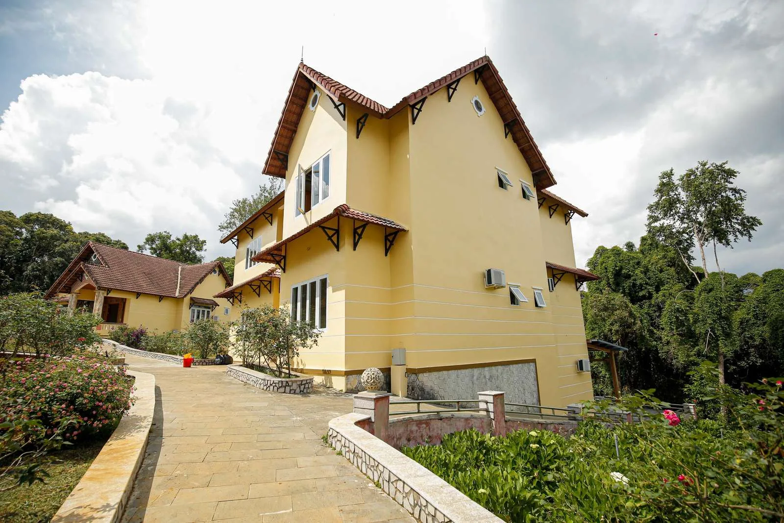 Villa sơn vàng mái ngói đỏ của Dakke Resort Măng Đen, lối đi lát đá dẫn qua vườn hoa, phía sau là cây xanh và trời nhiều mây