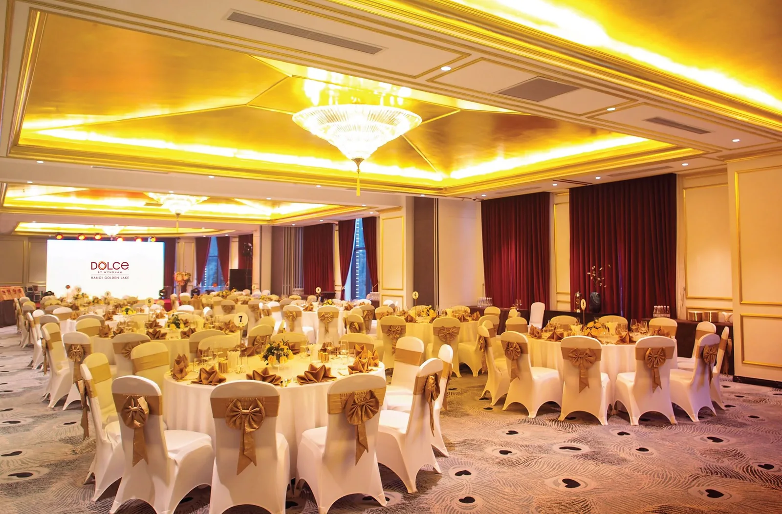Sảnh tiệc Dolce by Wyndham Hanoi Golden Lake xếp bàn tròn, ghế bọc nơ vàng, trần viền đèn vàng và màn chiếu phía cuối