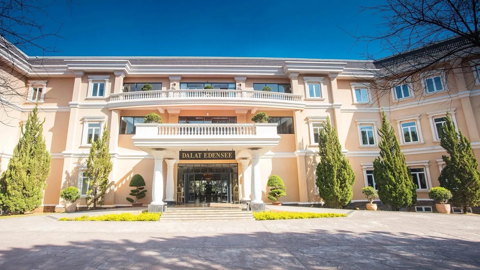 Tòa nhà chính Dalat Edensee màu hồng nhạt với hàng cột trắng, ban công và hàng cây tùng trước sảnh