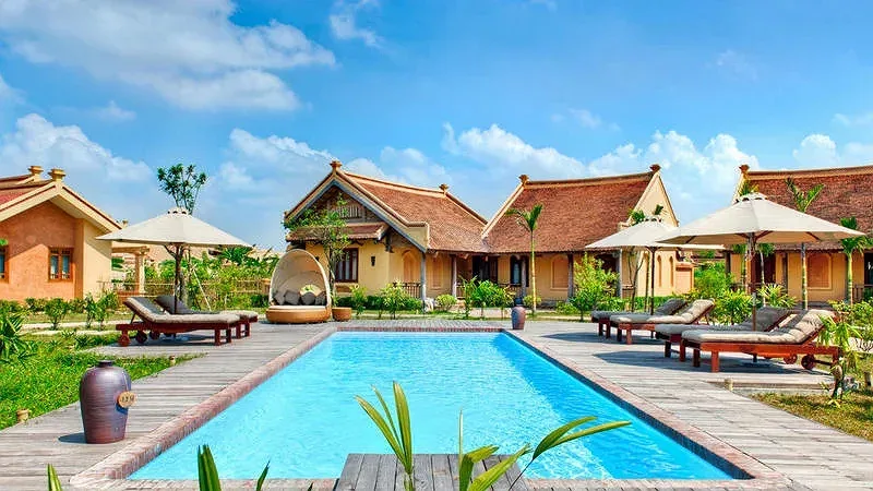 Dãy bungalow mái ngói nâu tường vàng quanh bể bơi ngoài trời, ghế tắm nắng và dù trắng tại Emeralda Resort Ninh Bình