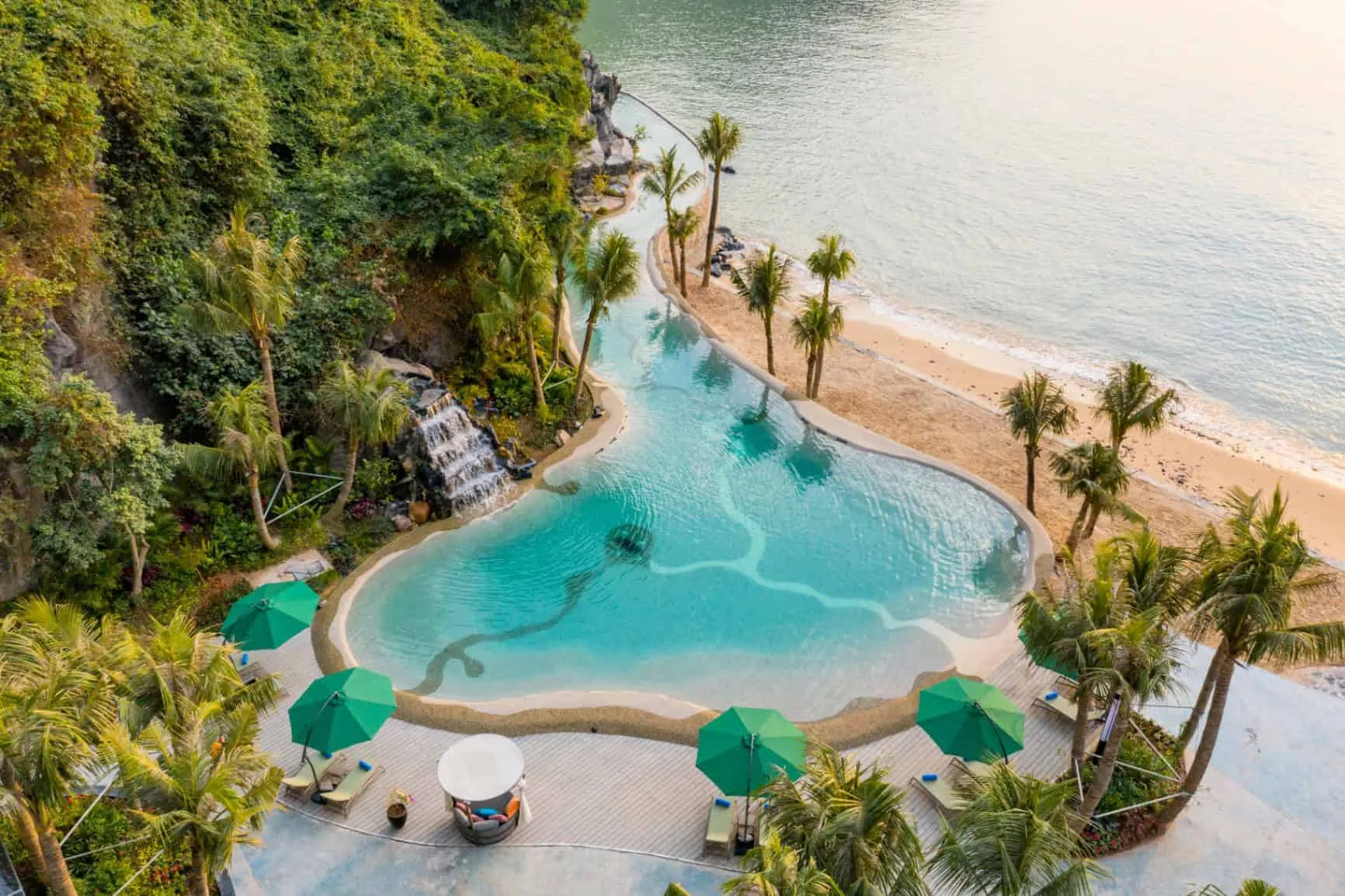 Charm Pool của Flamingo Cát Bà nhìn từ trên cao: bể uốn lượn có thác nước đổ từ vách đá, hàng dừa và dù xanh sát bãi cát