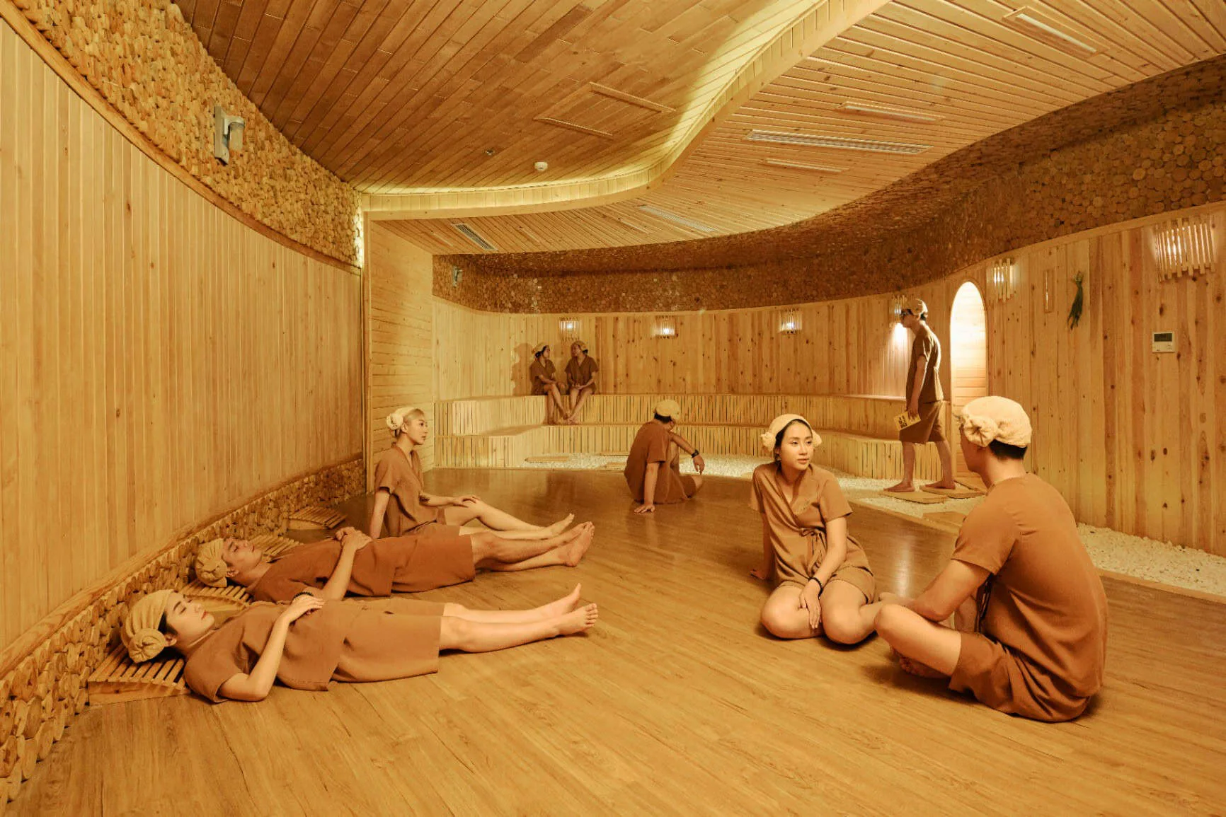 Khu xông Jjimjilbang ốp gỗ thông với trần lượn sóng tại SEVA Onsen & Spa, khách mặc đồ xông nằm ngồi thư giãn trên sàn gỗ