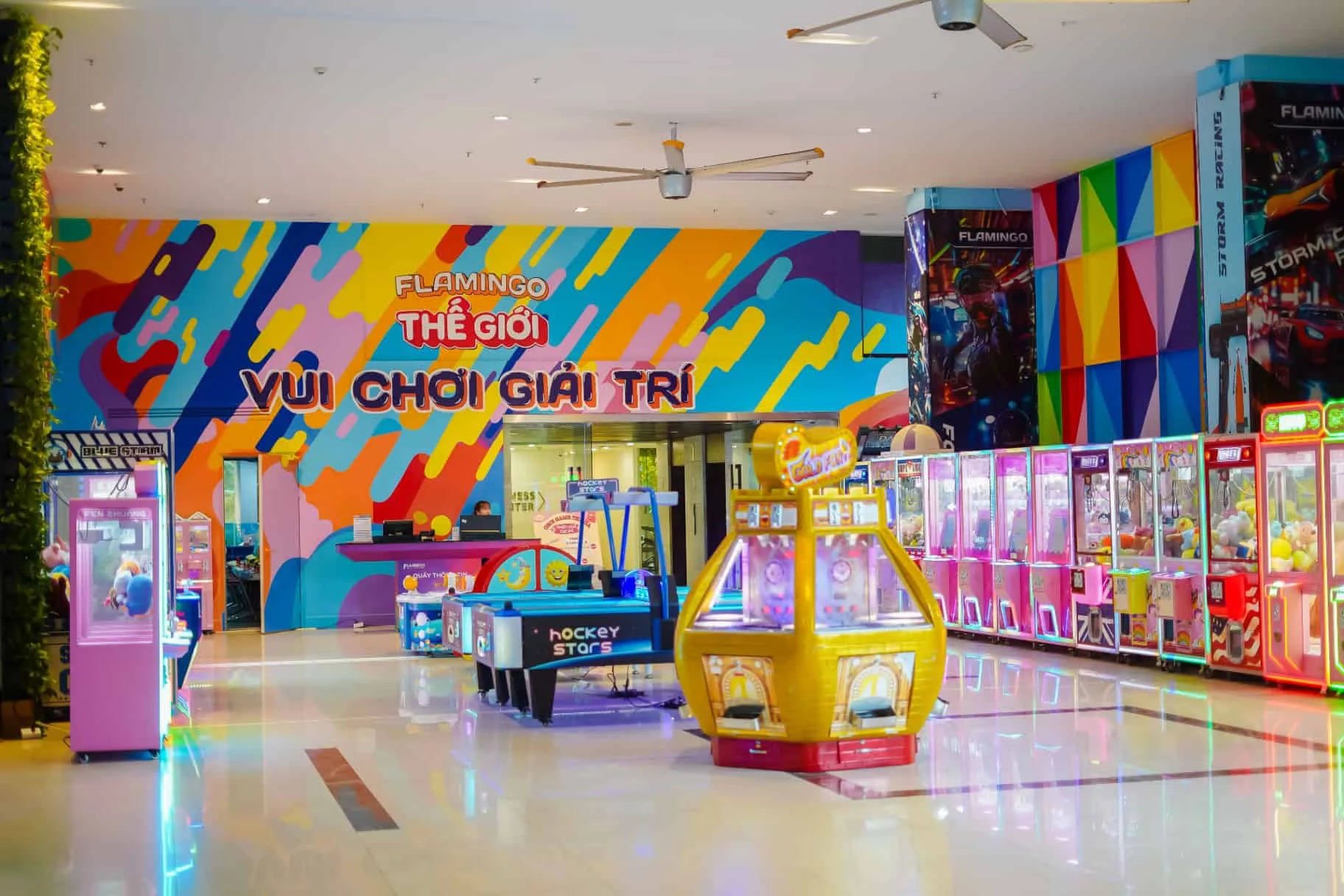 Khu Flamingo Play World trong nhà với tường graffiti chữ Thế giới vui chơi giải trí, bàn hockey và dãy máy gắp thú màu hồng