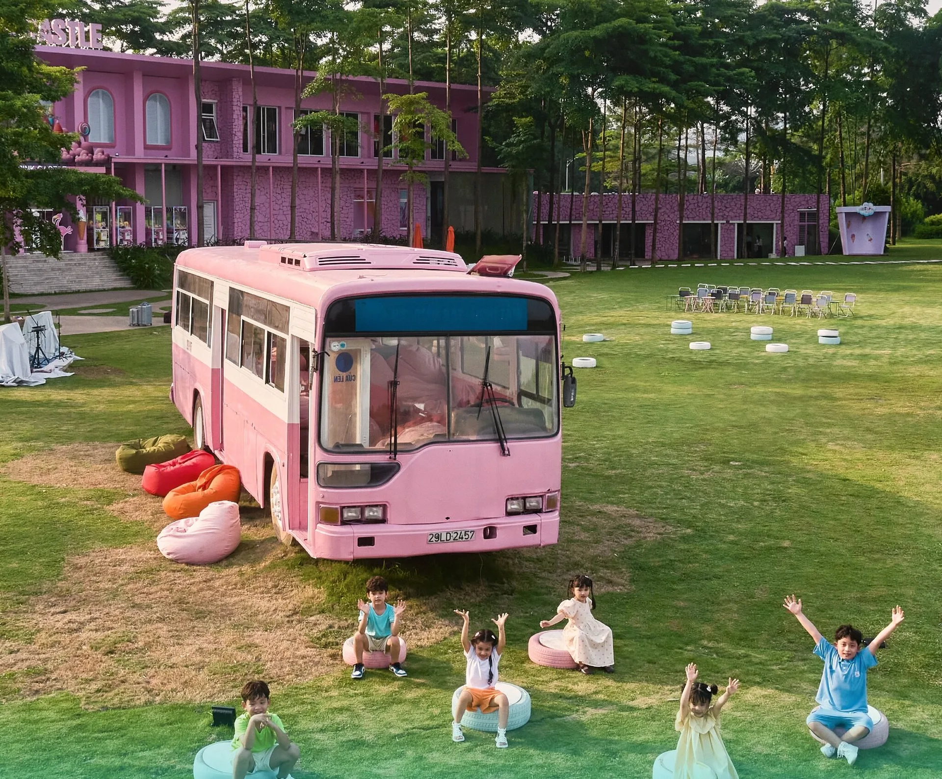 Nhóm trẻ em ngồi trên lốp xe sơn màu vẫy tay trên bãi cỏ trước chiếc xe buýt hồng và toà Pink Castle ở Flamingo Đại Lải
