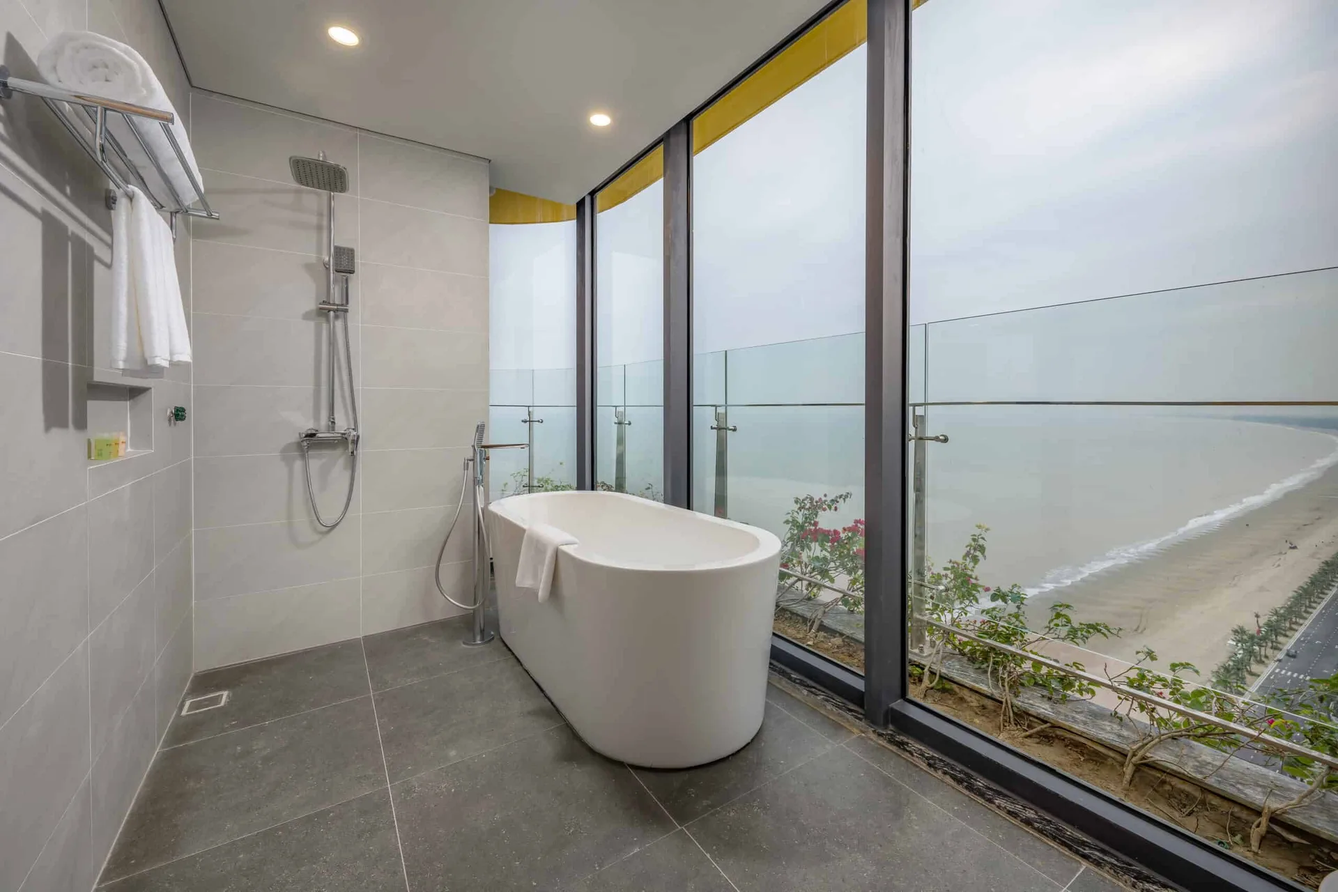 Phòng tắm căn Premier Residence Ocean View 02 phòng ngủ tại Flamingo Hải Tiến Resort, bồn tắm cạnh vách kính nhìn xuống bãi cát dài và đường ven biển trồng cọ