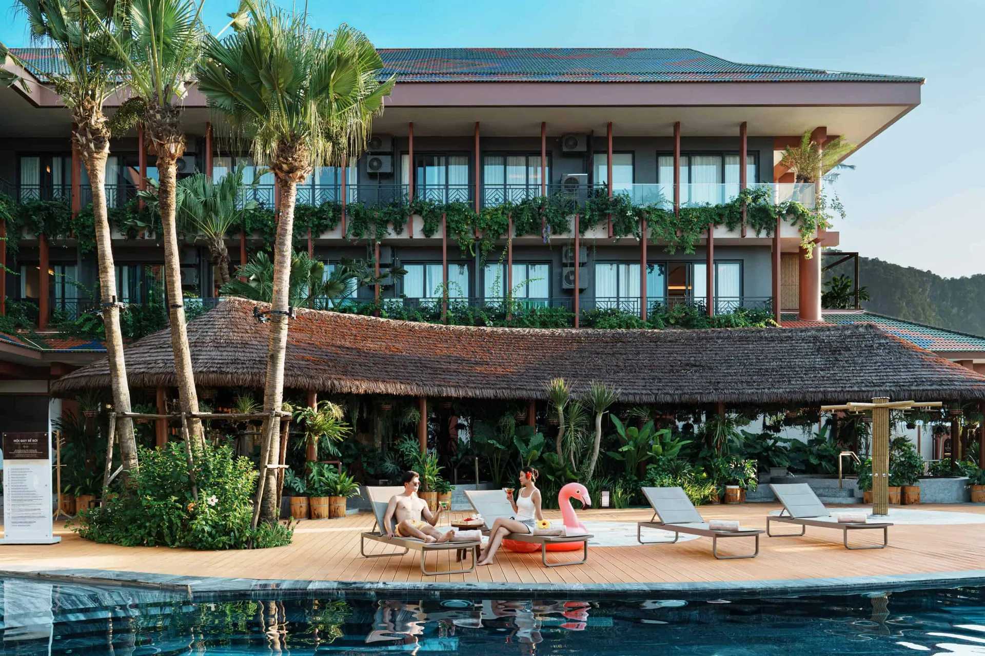 Bể bơi ngoài trời tại Flamingo Heritage Onsen & Resort Tân Trào, sàn gỗ có ghế tắm nắng và phao hồng hạc, tòa khách sạn 3 tầng phía sau