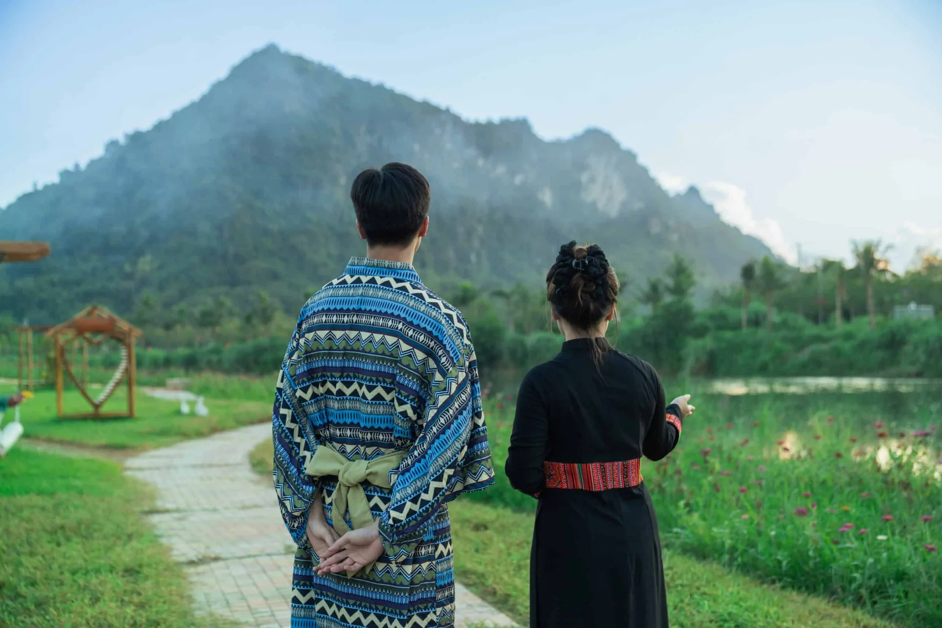 Hai người mặc trang phục hoa văn thổ cẩm đứng trên lối đi ven sông, nhìn về ngọn núi đá vôi mờ sương