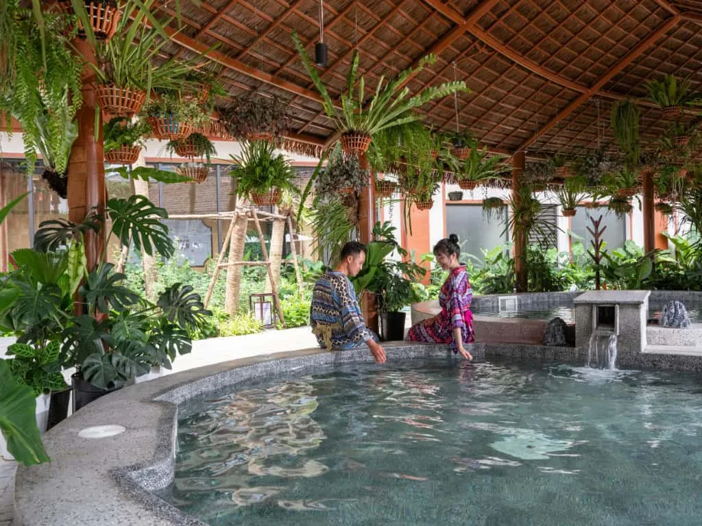 Bể khoáng nóng dưới mái lá treo nhiều chậu dương xỉ, hai khách mặc trang phục thổ cẩm ngâm chân tại Flamingo Heritage Onsen & Resort Tân Trào