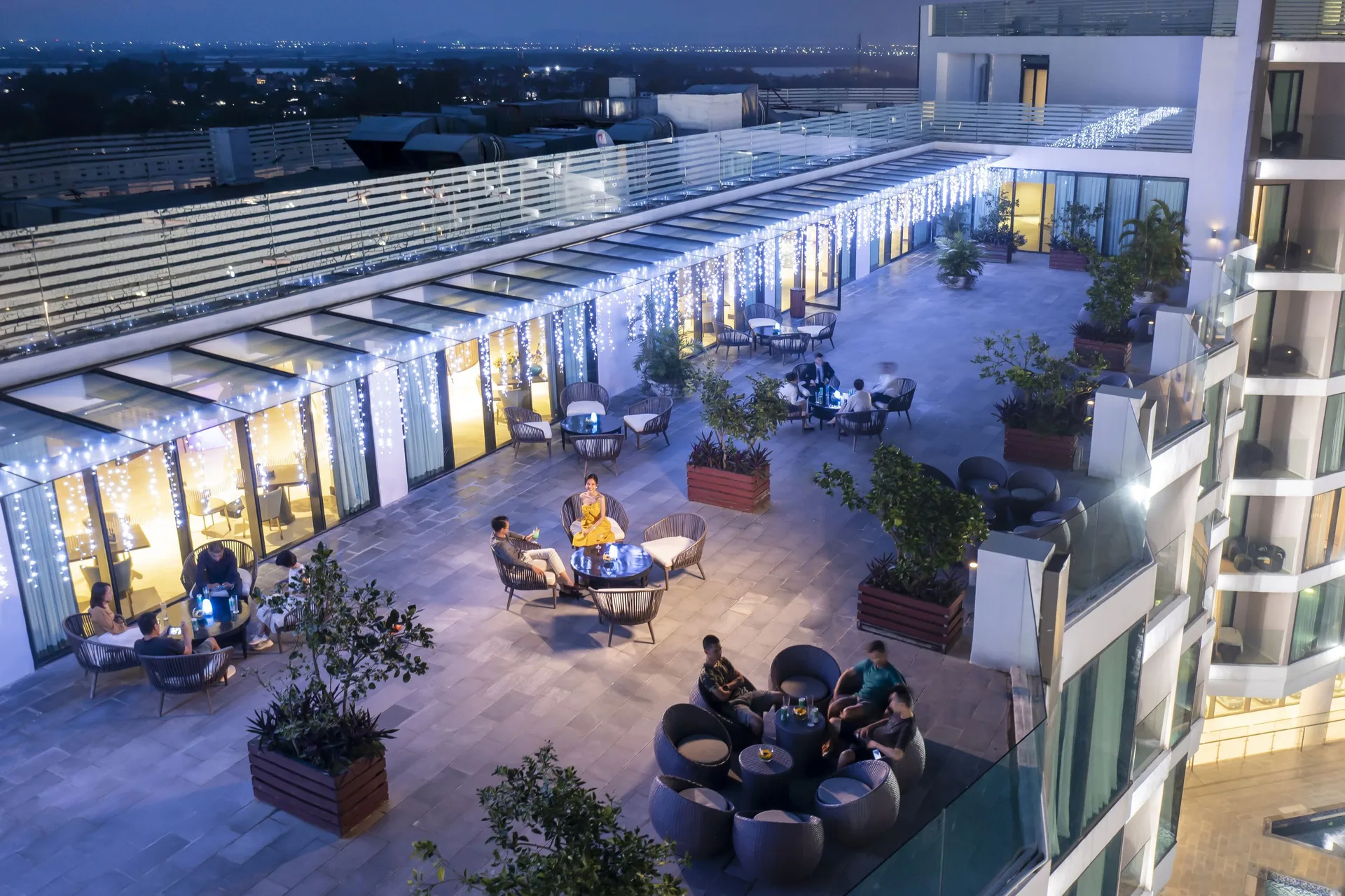 Sky Bar trên sân thượng FLC Grand Hotel Sầm Sơn buổi tối, dây đèn trang trí, các nhóm ghế mây quanh bàn thấp nhìn ra thành phố lên đèn