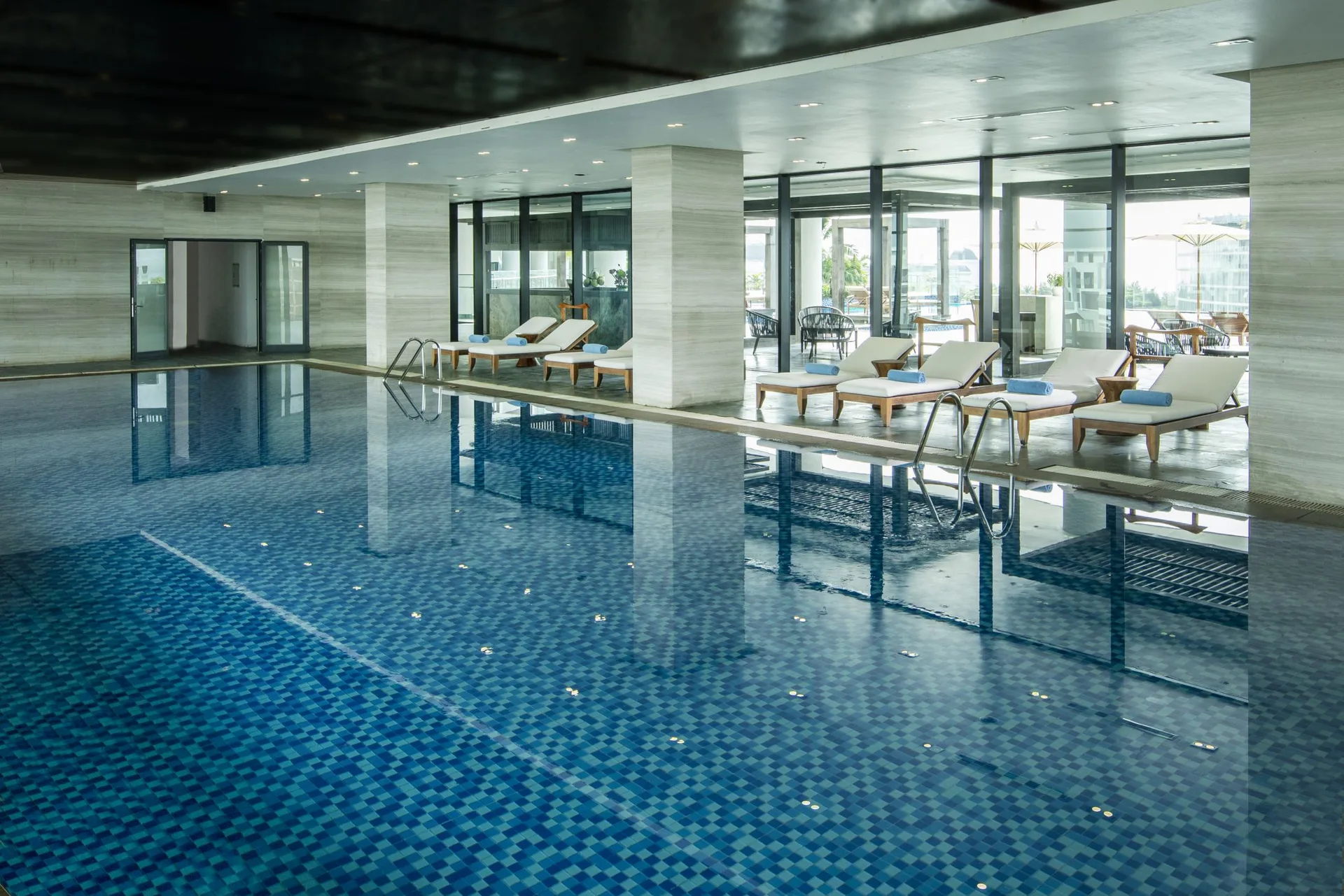 Bể bơi trong nhà lát gạch xanh của FLC Grand Hotel Sầm Sơn, hàng ghế tắm nắng và cửa kính mở ra sân ngoài trời