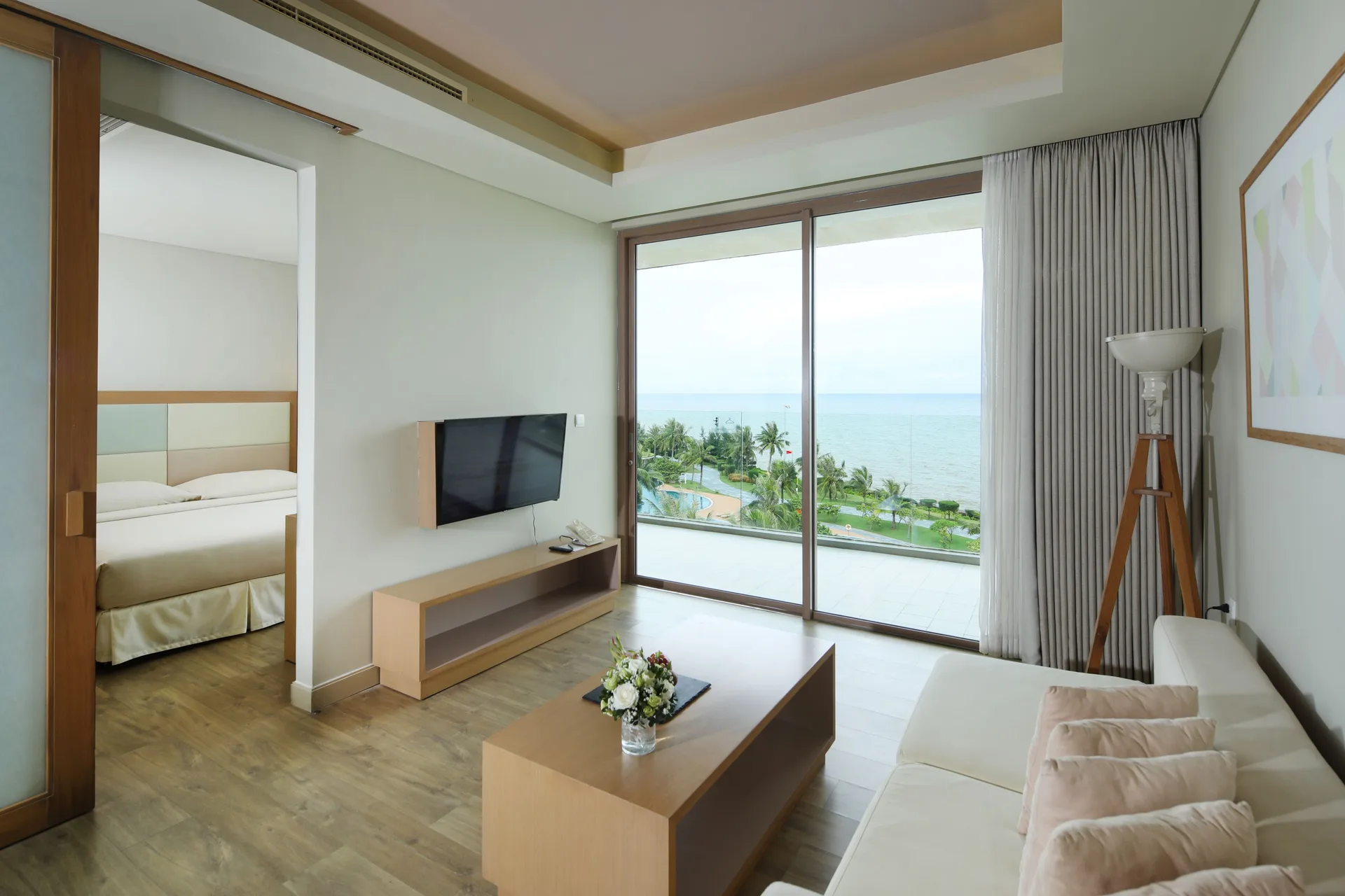 Phòng khách Family Suite với sofa kem, TV, cửa kính mở ra ban công nhìn bể bơi, hàng cọ và biển Sầm Sơn