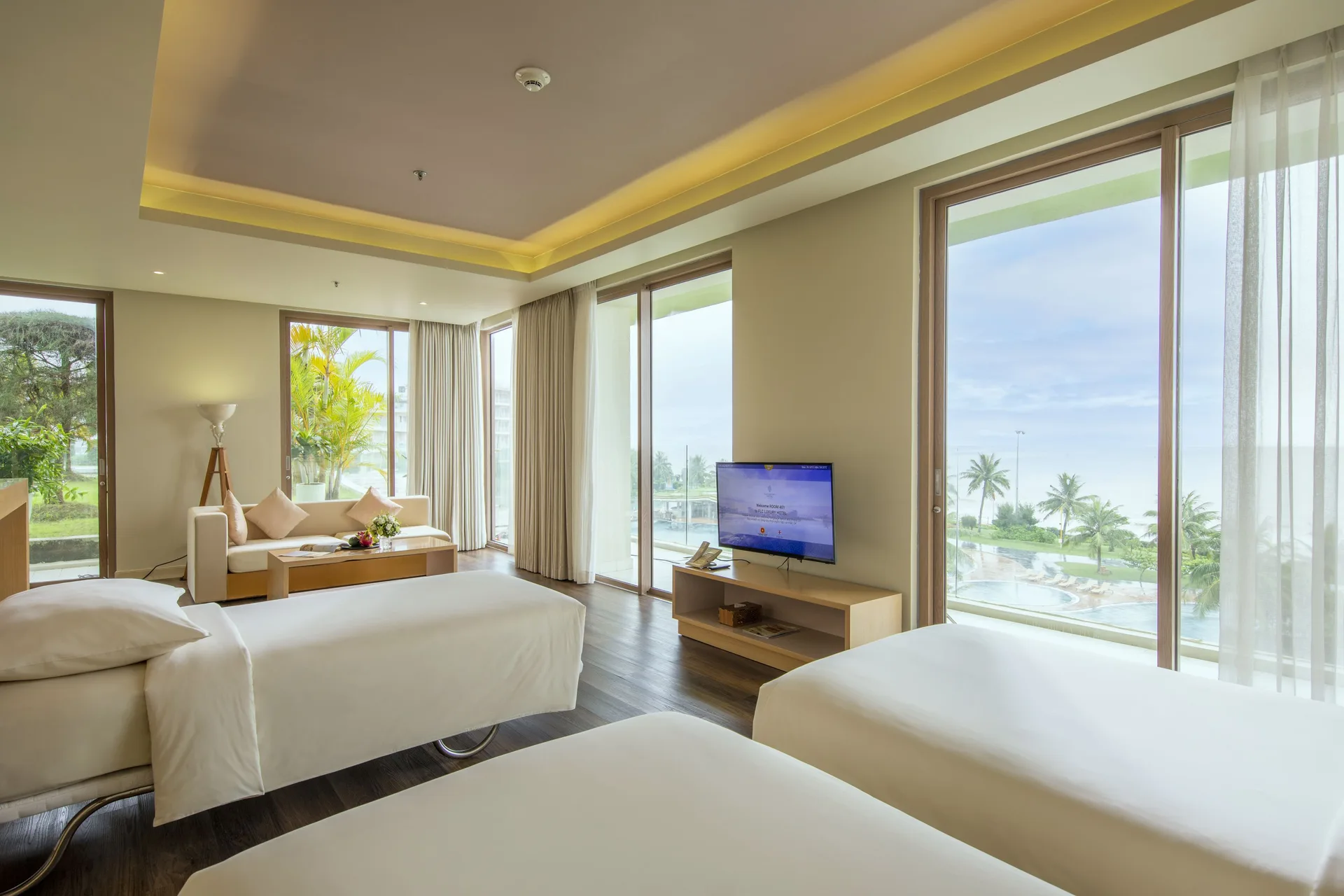 Studio Suite Twin bed của FLC Luxury Hotel Sầm Sơn: góc sofa, giường đơn và cửa kính lớn nhìn xuống bể bơi, hàng dừa và biển
