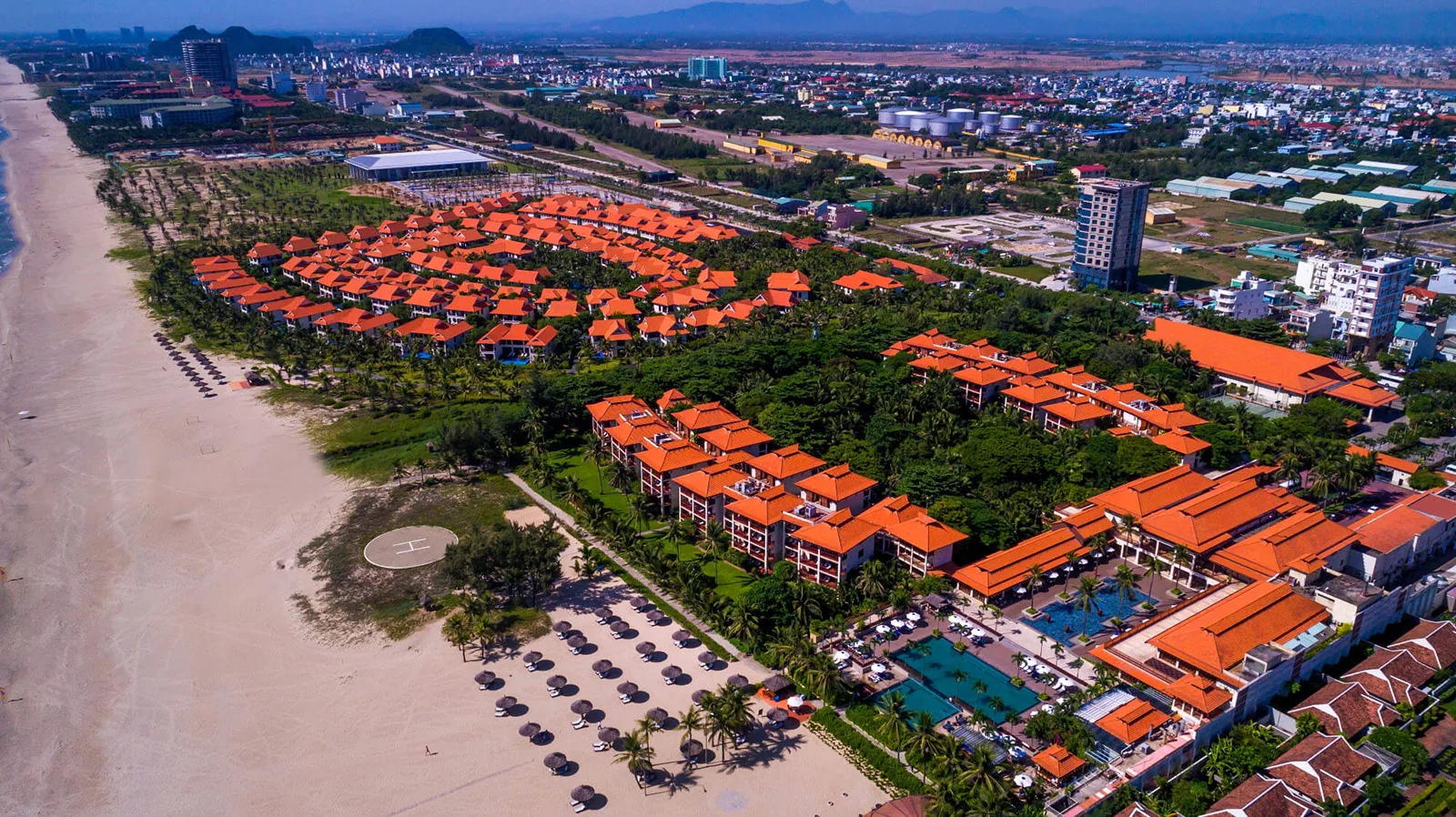 Furama Resort Danang nhìn từ trên cao: các dãy nhà mái ngói cam giữa vườn cây, hồ bơi và hàng dù trên bãi cát dài