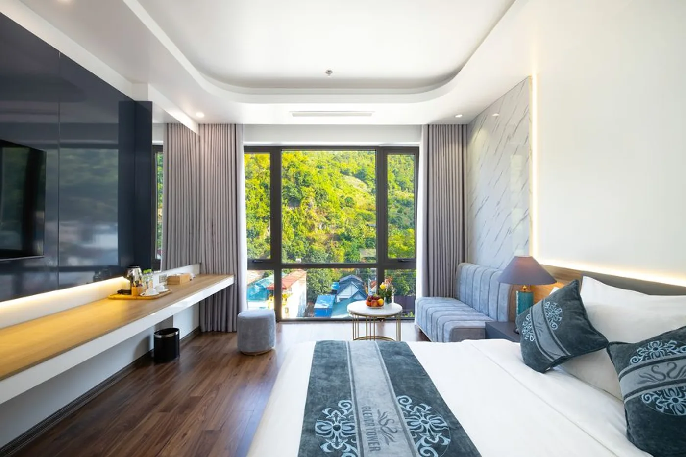 Phòng Deluxe Hướng Núi của Glenda Tower Mộc Châu Hotel với giường trắng, sofa xám và khung cửa kính lớn nhìn ra sườn núi xanh