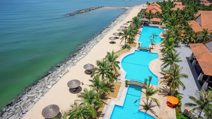 Ảnh từ trên cao hồ bơi nhiều tầng nối cầu của Golden Sand Resort & Spa Hoi An, bên trái là bãi cát có dù lá và biển xanh