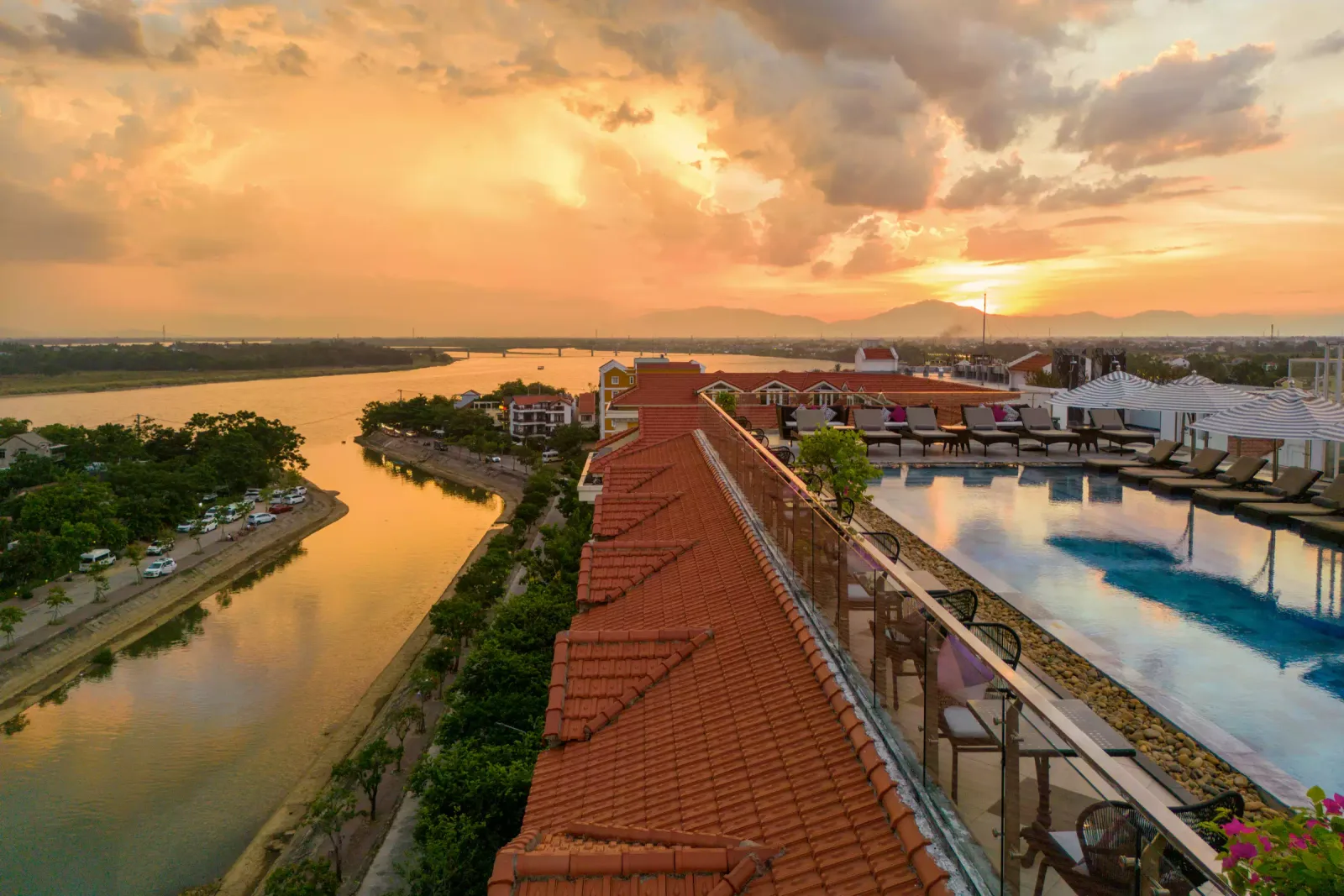 Bể bơi tầng thượng của Hotel Royal Hoi An - MGallery nhìn xuống sông Thu Bồn lúc hoàng hôn, mái ngói đỏ và hàng ghế nằm bên phải
