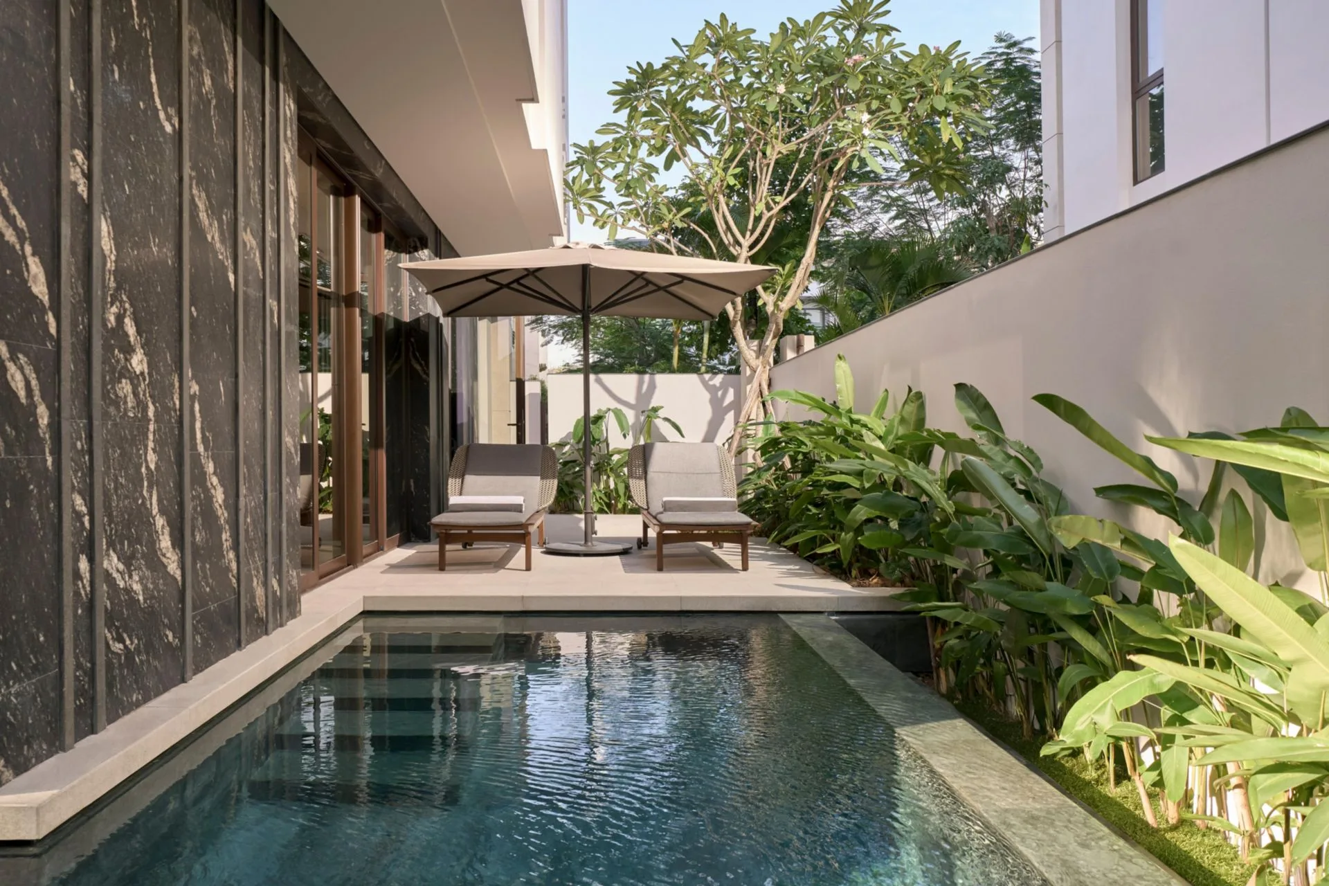 Hồ bơi riêng của một biệt thự, hai ghế tắm nắng dưới ô dù, tường đá đen vân trắng và hàng chuối cảnh