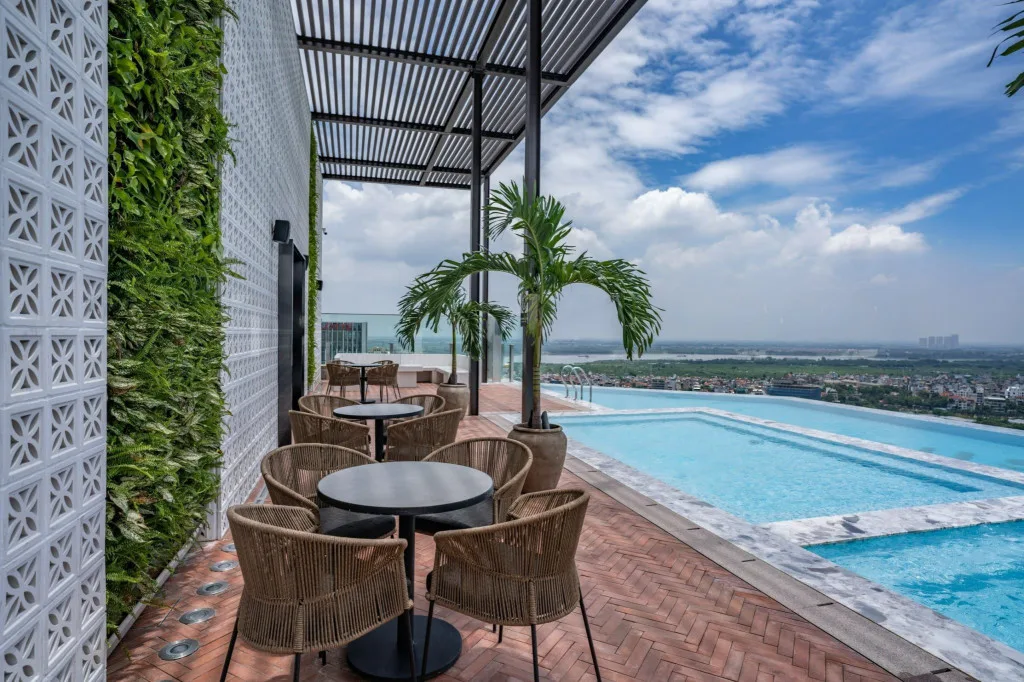 Bể bơi vô cực tầng thượng L7 West Lake Hanoi by Lotte cạnh bàn ghế mây dưới giàn che, tường cây xanh, nhìn ra toàn cảnh thành phố