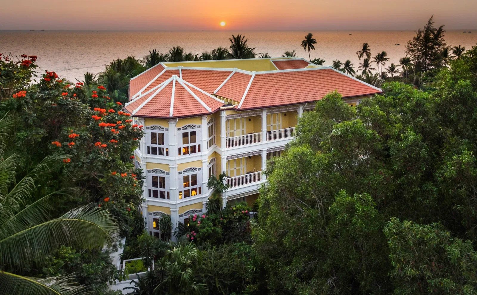 Tòa nhà tường vàng mái ngói đỏ của La Veranda Resort Phu Quoc - MGallery giữa rừng cây, phía sau là biển lúc mặt trời lặn