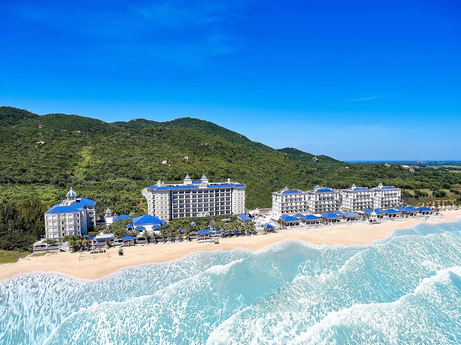 Toàn cảnh Lan Rừng Resort & Spa Phước Hải với các tòa nhà mái xanh cobalt nằm dọc bãi cát, sóng trắng phía trước và đồi xanh phía sau