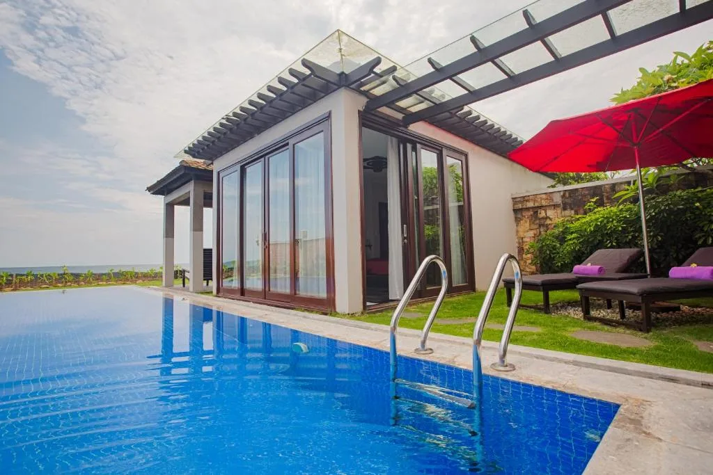 Pool Villa ở Lapochine Beach Resort (Ana Mandara Huế): hồ bơi riêng xanh trước nhà cửa kính, ghế nằm và ô đỏ trên bãi cỏ, biển phía xa