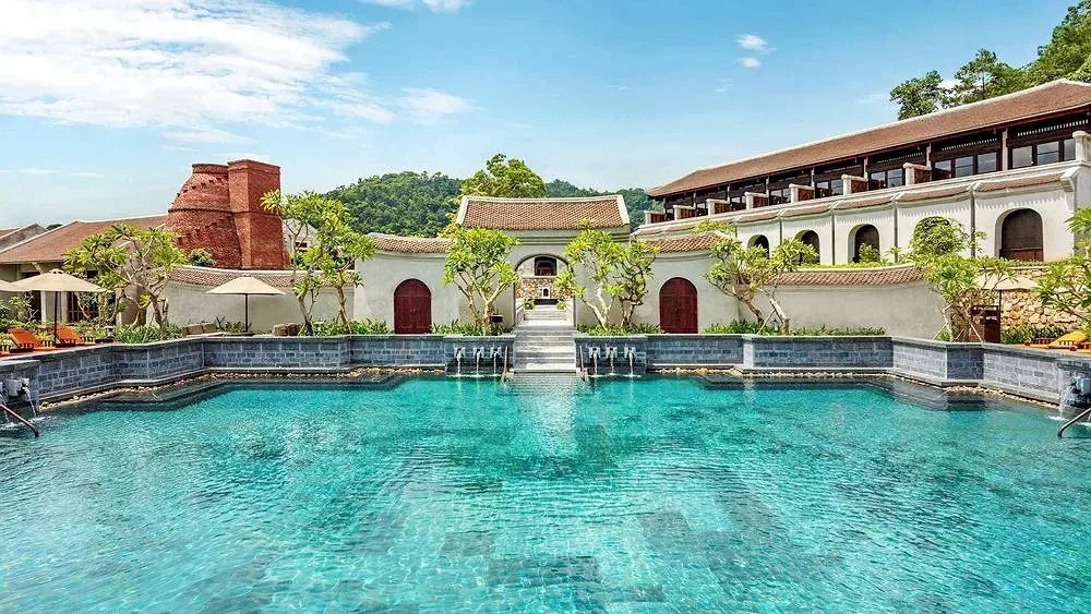 Bể bơi khoáng nóng Zen Pool của Legacy Yên Tử, nước xanh ngọc giữa tường trắng cửa vòm đỏ và dãy nhà mái ngói, phía sau là đồi xanh