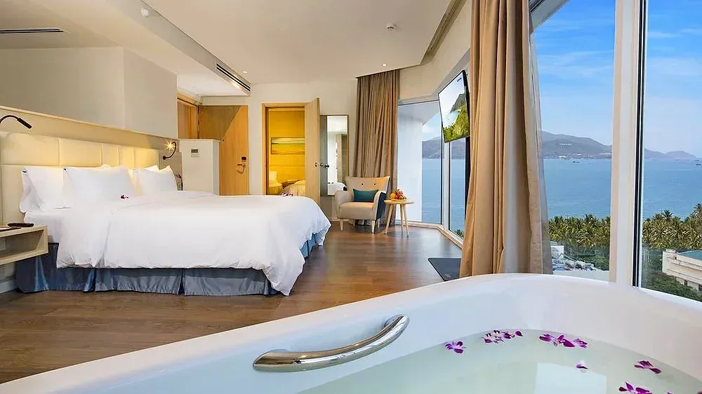 Bồn tắm rải hoa lan tím đặt cạnh giường, cửa kính lớn nhìn ra vịnh Nha Trang trong phòng Executive Ocean View tại Liberty Central