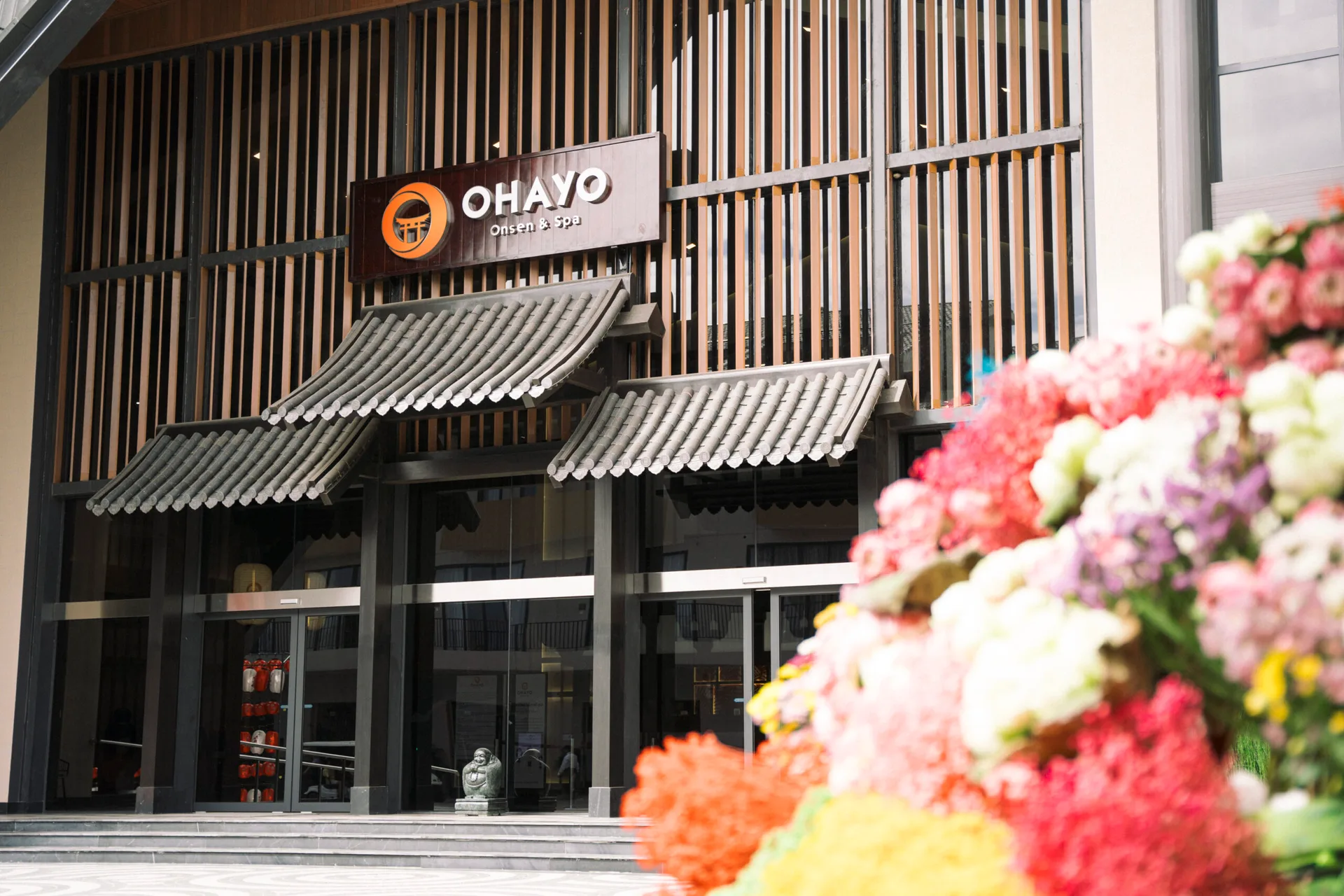 Lối vào Ohayo Onsen & Spa tại Lynntimes Thanh Thủy với biển hiệu logo cam, mái ngói kiểu Nhật trên cửa kính và hoa nhiều màu ở tiền cảnh