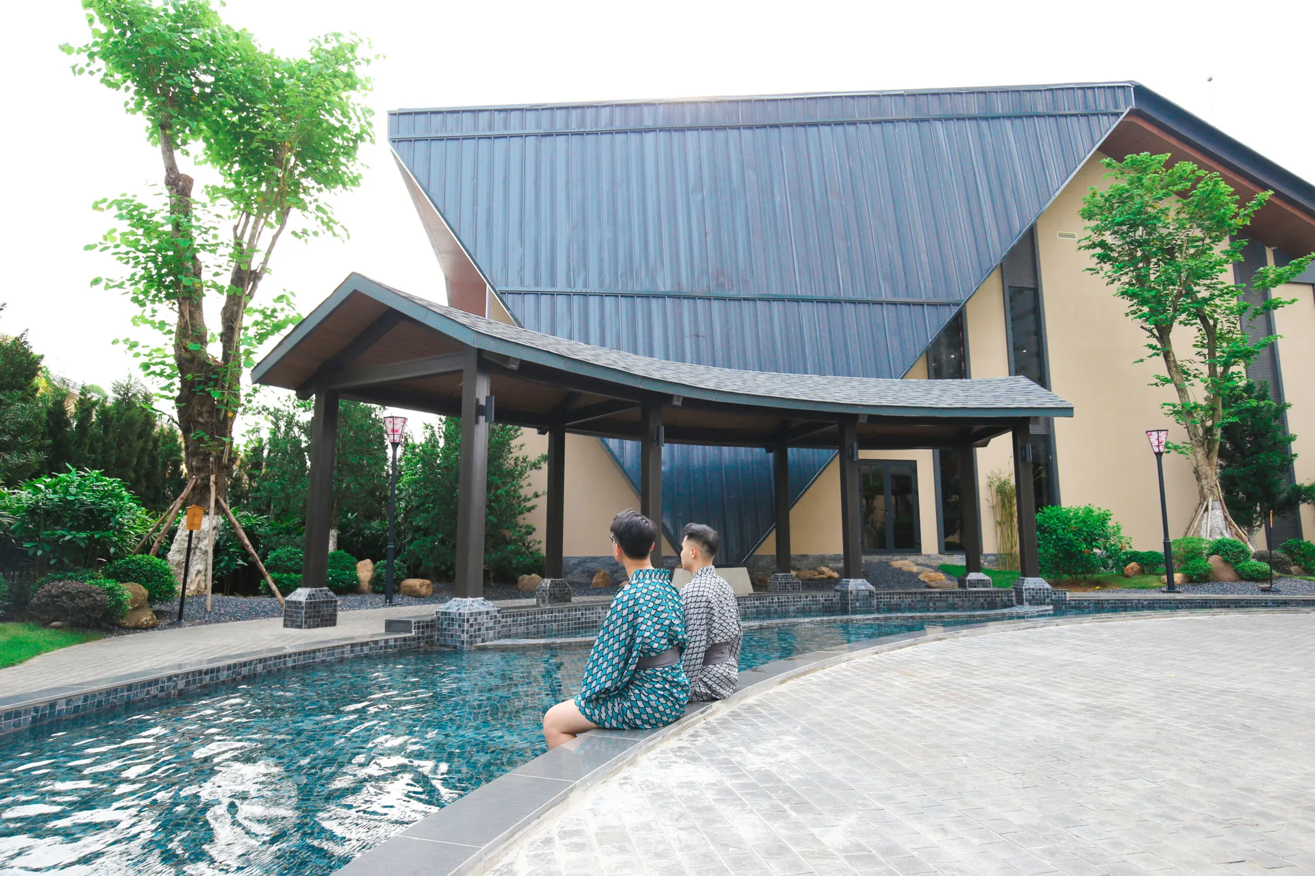 Hai khách mặc yukata ngồi ngâm chân bên bể khoáng ngoài trời lát đá xanh, chòi mái cong kiểu Nhật và tòa nhà Ohayo mái xanh đen phía sau