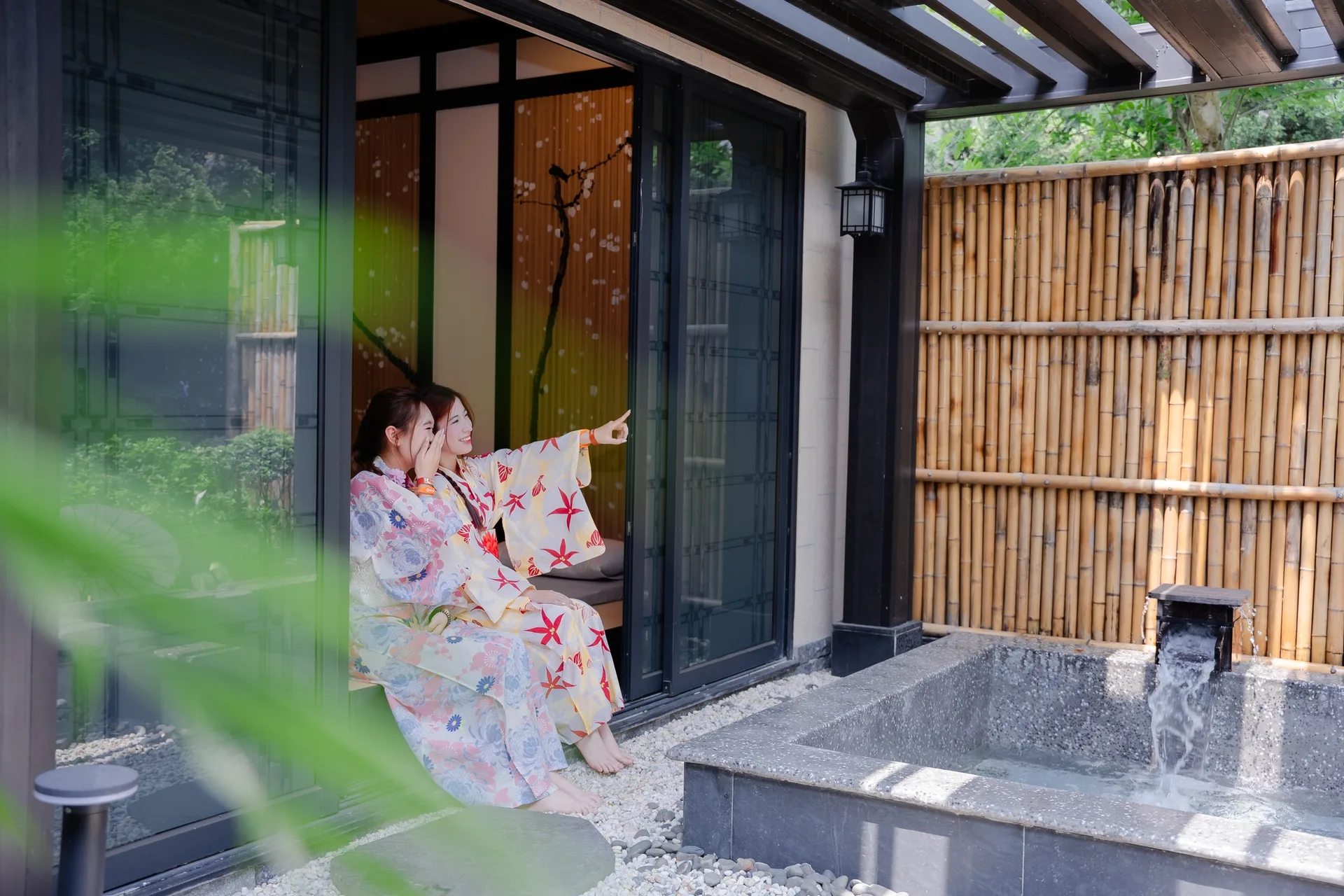 Phòng Private Onsen: hai khách mặc yukata ngồi ở hiên phòng tatami, bên cạnh là bồn khoáng đá vuông có vòi xả nước và tường rào tre