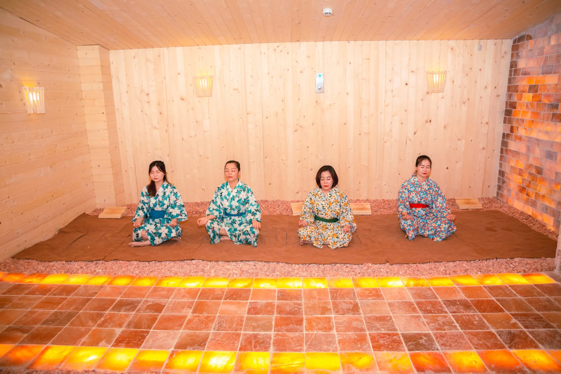 Phòng xông đá muối Himalaya trong Ohayo Sauna, tường gạch muối phát sáng màu cam, bốn khách mặc yukata ngồi thiền trên sàn rải đá muối