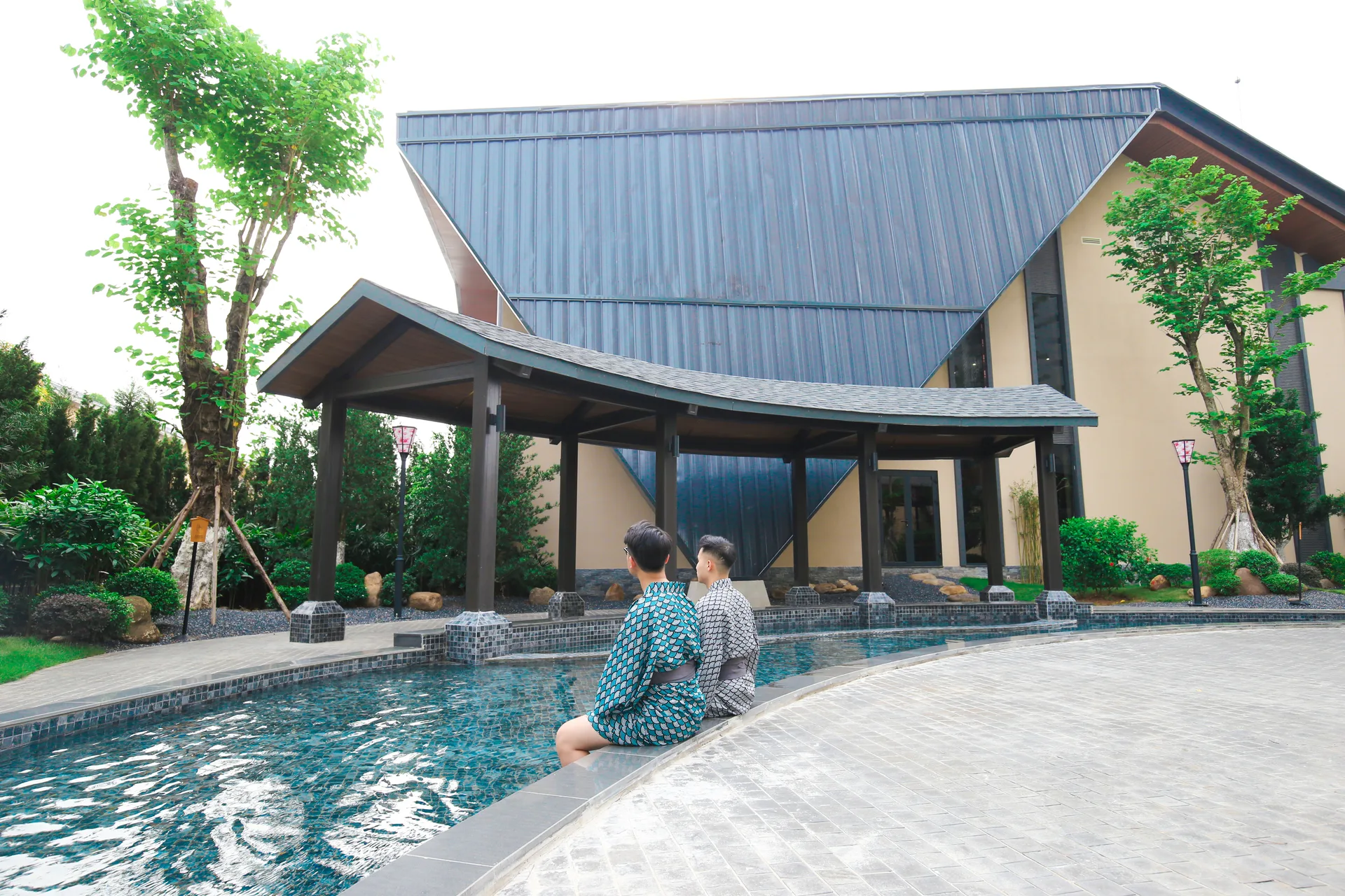 Hai khách mặc yukata ngồi ngâm chân ở bể khoáng ngoài trời trước tòa Ohayo Onsen & Spa của Lynntimes Thanh Thủy