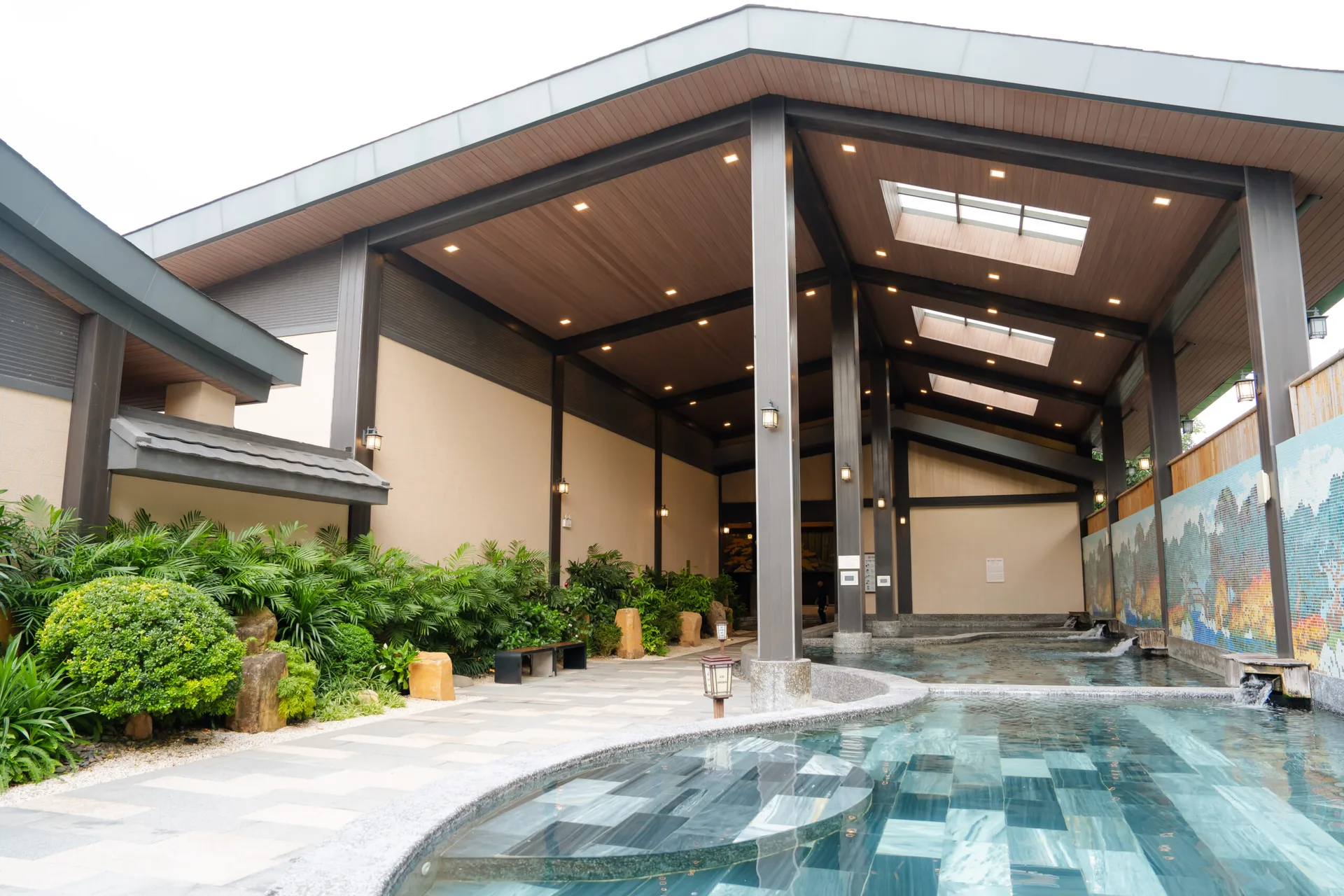 Bể Natural Onsen ngoài trời uốn lượn dưới mái gỗ lớn ở Lynntimes Thanh Thủy, tường mosaic phong cảnh và vòi gỗ chảy nước