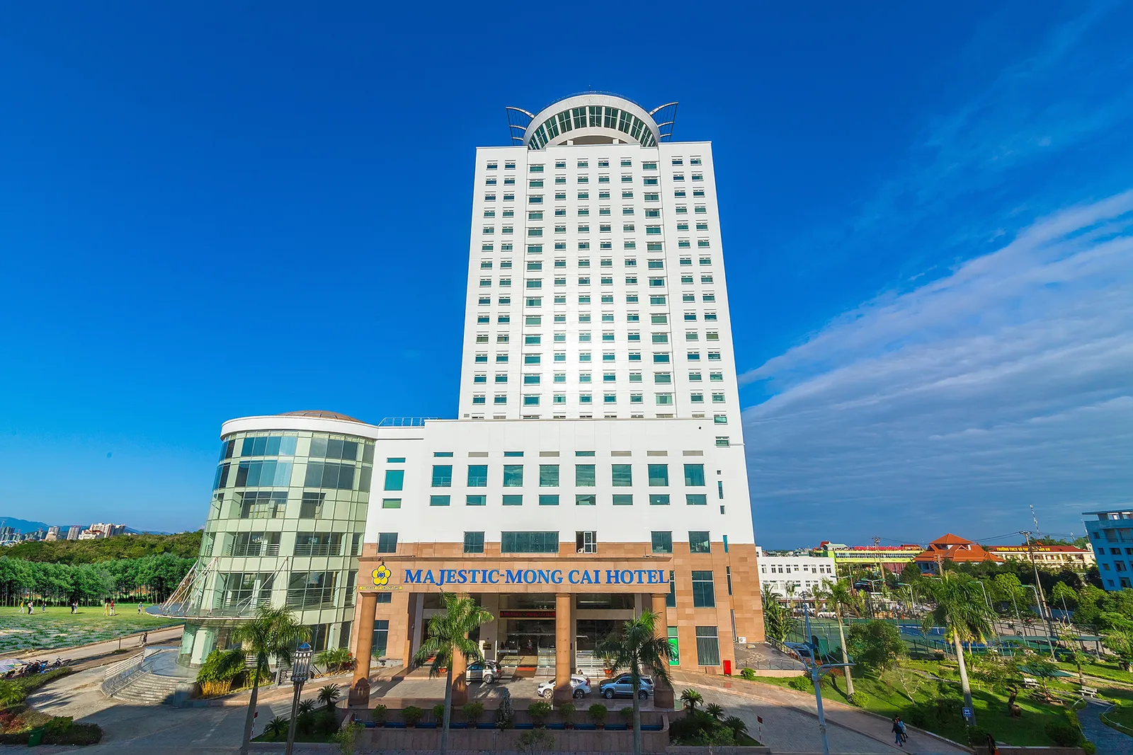 Majestic Móng Cái Hotel, toà tháp trắng có vòm kính trên đỉnh, sảnh chính gắn biển tên khách sạn dưới trời xanh