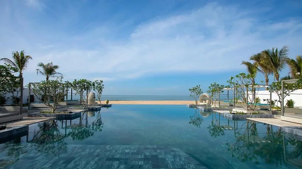 Bể bơi vô cực của Melia Hồ Tràm Beach Resort, mặt nước phản chiếu cây xanh, phía cuối là bãi cát và biển