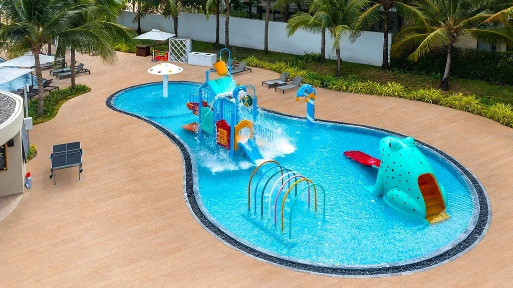 Hồ nước trẻ em tại Melia Hồ Tràm Beach Resort nhìn từ trên cao, có cầu trượt nhiều màu, vòng phun nước và mô hình chú ếch xanh giữa sàn gỗ và hàng dừa