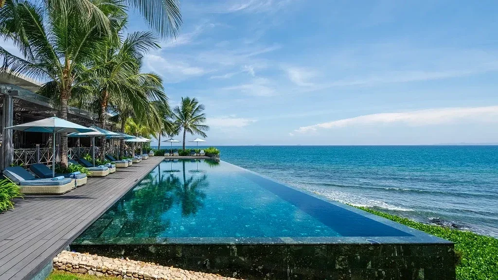 Hồ bơi vô cực của Mia Resort Nha Trang chạy dọc mép biển, hàng dừa và ghế tắm nắng bên sàn gỗ