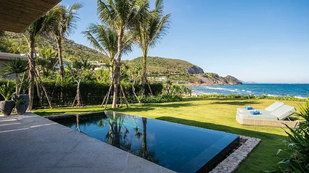 Hồ bơi riêng tràn bờ trước villa Mia Resort Nha Trang, bãi cỏ, ghế tắm nắng, hàng dừa và biển xanh dưới chân núi