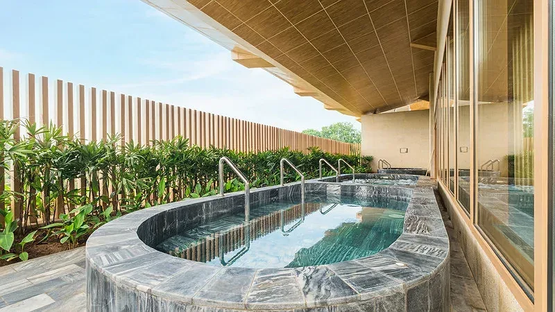 Bồn ngâm khoáng nóng ốp đá xám ngoài trời tại Minera Hot Springs Bình Châu, có tay vịn inox, hàng cây xanh và vách gỗ bao quanh