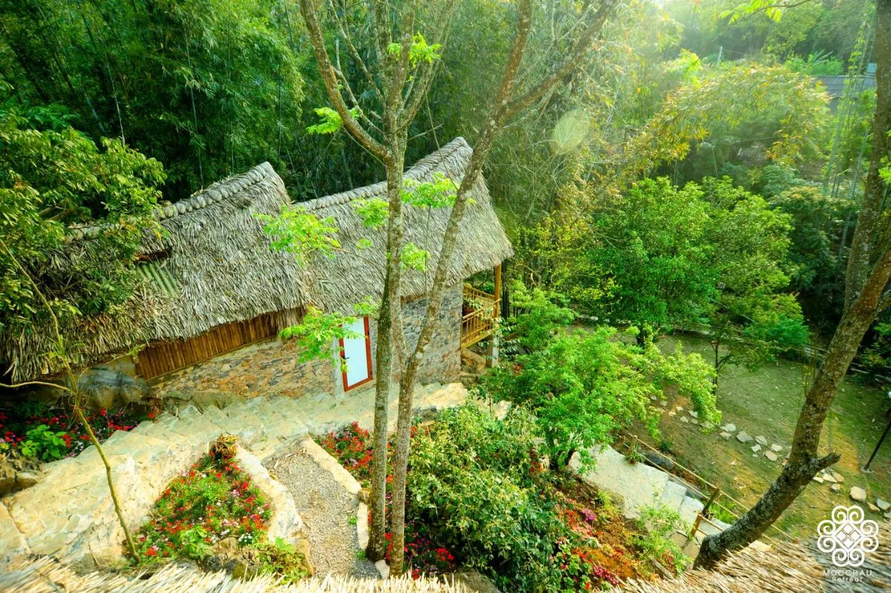 Bungalow tường đá mái lá cọ của Mộc Châu Retreat giữa vườn cây, bậc đá dẫn xuống có hoa đỏ hai bên